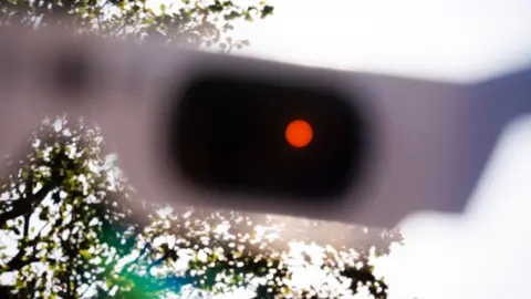 PA Media The sun looks orange, polarised through a pair of solar eclipse glasses with a tree behind.