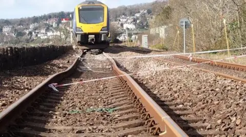 RAIB Looking up from train tracks to a carriage which has derailed on its way to Barrow-in-Furness, in Cumbria. The train is Northern operated, with blue and yellow branding. The train tracks have collapsed into a hole in the ground.