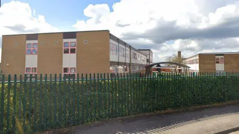 Google The school building seen from the side, behind a long green metal fence. There are two blocks from brown brick with long lines of windows and a green space between them. 