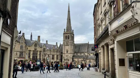 Saddler Street in the City of Durham. The road contains old buildings and opens out into a square, at the end of which is a church with a tall steeple. Several people are walking around the square.