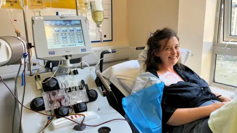 PA Media Jessica Kent-Hazledine sits on a hospital bed while receiving a plasma transfer. A large machine is next to her with wires sticking out of it. Kent-Hazledine has a black blouse on which is partially open to allow the treatment to be carried out. She has a broad smile on her face.