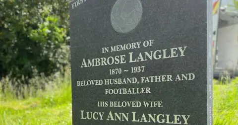 A close-up of a dark grey granite headstone with the wording, 'In memory of Ambrose Langley 1870 - 1937 beloved husband, father and footballer, his beloved wife Lucy Ann Langley'