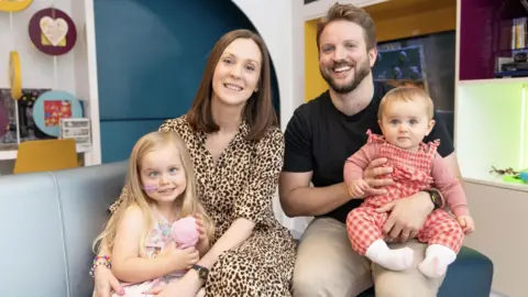 UCLH The family pose for a happy picture in the hospital play room. Clare has long brown hair and wears a leopard print dress. Her arms are around Seren, whose eyes look slightly misaligned here. Mark has short brown hair and a beard and is holding their other daughter, a little baby with fair hair wearing a red-checked all in one outfit.