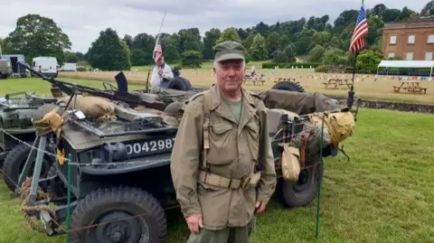 Dudley Council A man in green and brown military clothing is standing in front of a military vehicle that is covered in brown sandbags and black guns. He is wearing a green hat and there are American flags on the vehicle.