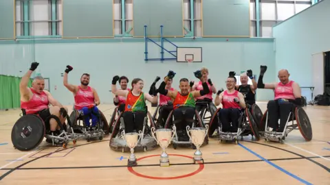 The Penrith Pumas wheelchair rugby team. They are inside a sports hall and posing in a group formation, with three people in the front row and seven behind them. The players are a mixture of men and women and they are cheering with their fists or arms on the air.