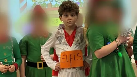 Supplied A young boy on stage wearing an Elvis Presley costume. An orange sign is hung over him reading "Elfis" 