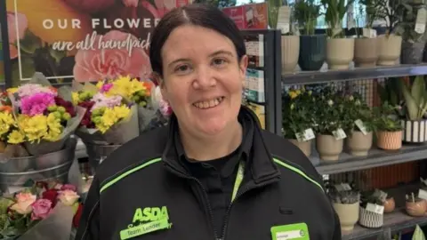 Woman in Asda uniform with dark hair smiles at the camera inside a supermarket.