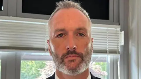 Det Con Lee Harshey-Jones. He looks at the camera with a neutral expression, and wears a white shirt, black suit jacket and bronze tie. He has short grey hair and short stubble.