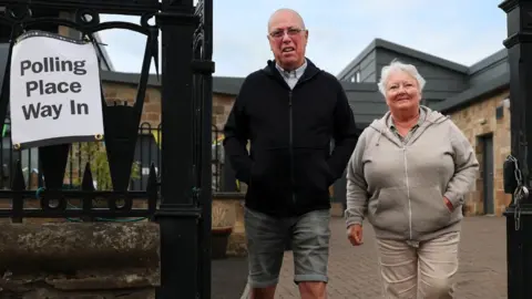 A couple, a bald man and a grey-haired woman, walk through gates next to a polling place sign. A stone building can be seen behind them 