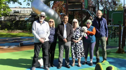 Six people and a dog standing in front of a new slide and a multi-coloured surface.