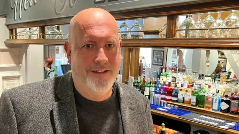 Landlord Neil Taylor, standing in front of his bar, wearing a jumper and blazer. There's a row of upturned glasses behind him as well as rows of colourful gin bottles. Customers can be seen in a mirror reflection, which is behind the bar.