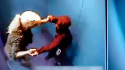 A still from CCTV footage showing a woman with blonde hair in a ponytail grabbing the wrists of a boy wearing a red hoodie as she pushes him into a windowless room with blue padding on all of the walls