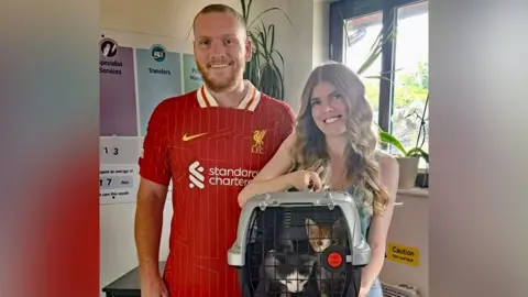Cats Protection Bradley Harding and Megan Green collect Fish and Chips from the rescue centre. Mr Harding is wearing a red sports top and Ms green is wearing a summer dress. She is holding the two kittens are in a cat carrier.