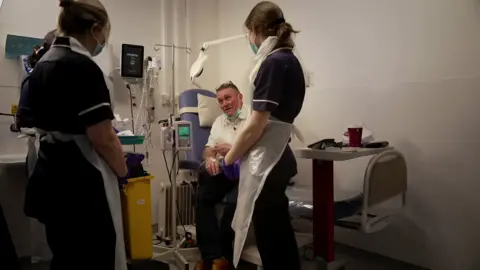 Whatney talks to nurses in a ward whilst hooked up to a machine. The nurses stand around him listening