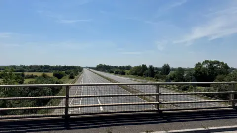 BBC/Dean Poolman Picture from the motorway bridge of an empty road and motorway. There is blue skies in the background.
