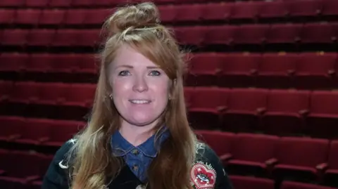 Shaun Whitmore/BBC Hannah Houghton is standing in the New Wolsey theatre behind her are rows of empty theater seats. The seats are a rich red colour and all the seats flipped up as they are empty. Hannah is wearing a blue denim shirt under a black patterned cardigan. Hannah has a top knot in her hair and is smiling at the camera 
