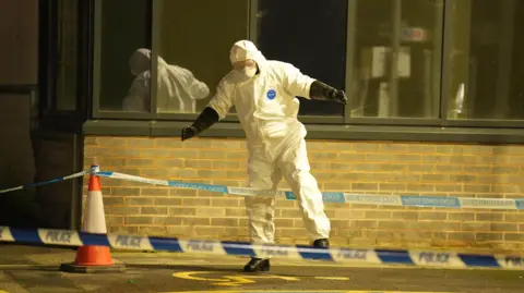 A man in a protective white suit. He is stepping over police tape that has cordoned off an area.