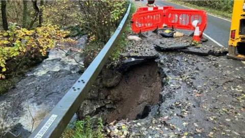 A hole can be seen in the A470 in Talerddig, with barriers around it and the river seen flowing below, with trees either side.