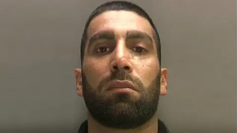 Mugshot of a man with a black beard and hair with a scar on his nose