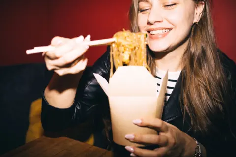 Getty Images A young woman uses chopsticks to lift noodles out of a takeaway box.