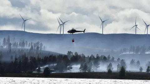 A helicopter hovers over heather, dropping water on a fire. The background is a hill with five wind turbines
