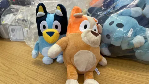 Two soft toys, based on dog characters, rest on a table in front of a bag of similar toys and counterfeit headphones.