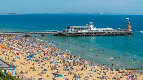 Bournemouth beach, a sandy beach which is packed with hundreds of people, tents and umbrellas. Bournemouth pier is in the background.