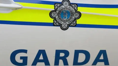 The image depicts the side of a white An Garda Síochána, Irish police force, car with the words garda written in blue capital letters. Above the word are blue and yellow lines with the crest of the organisation also visible in the middle of the lines.