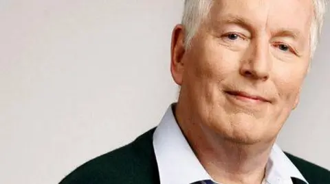 Richard Nankivell stands in front of a white screen. He has white grey hair and blue eyes. He is wearing a white shirt and a black jumper.