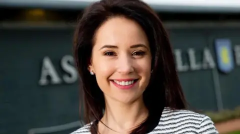 A woman with dark hair and a black and white top is smiling at the camera. The Aston Villa club crest is blurred in the background.