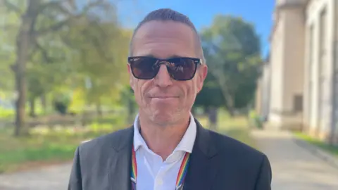 A man wearing sunglasses in a white shirt and dark grey jacket with trees and a university building in the background