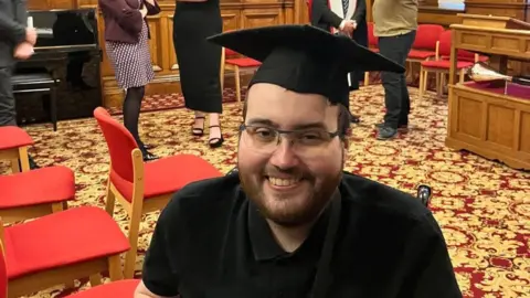 Supplied Daniel Swales, in a black t-shirt and with a black graduation cap on, is smiling at the camera. He has brown beard and is in a hall with red chairs.