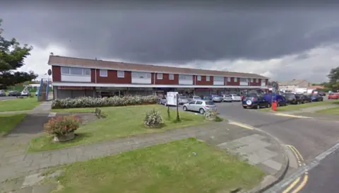 The shopping parade on Elizabeth Way. It is a row of ground floor shops with flats above. There are parked cars outside and a small green area before the junction with the road.