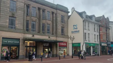 Stafford Borough Council A row of grey shops with signs reading The Body Shop and F Hinds on the ground floor. There is a sign reading Guildhall Shopping centre half-way up one wall. A few people mill around in front of the stores.