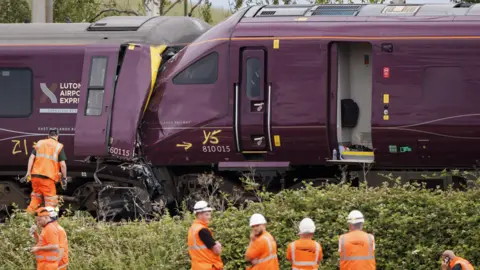 Two trains are seen crashed together with people wearing orange protective clothing standing alongside.
