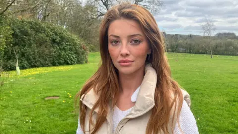 A woman with strawberry blonde hair stares at the camera. She's stood at a gate looking over a green field behind her, with hedges and trees in the background. The sky has grey clouds with a hint of blue skies in the distance. Lorna is wearing a white top, with a cream gillet over the top.