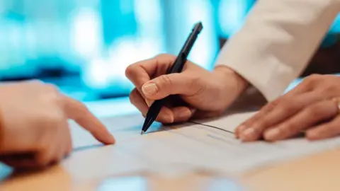 Getty Images Someone is signing a document with a black pen, only their hands are in the shot. They are wearing a white long sleeved piece of clothing. There is a another hand pointing to a point in the document.