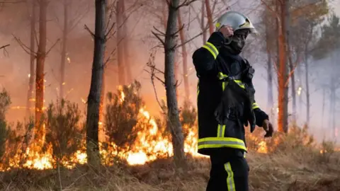 A firefighter stands in protective equipment in front of a wall of orange flames burning inside a forest. Picture date: 27 July 2026. 