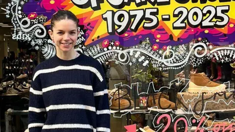 Jake Shoes Maisy Bland had dark hair tied back in a pony tail and is wearing a blue and white striped jumper, She is standing at the front of the shop which has 1975-2025 painted on the window in white and surrounded by yellow and purple.