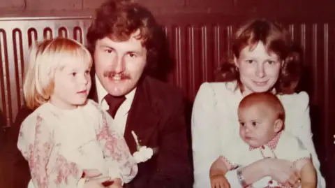 Scowen family Black and White photo of Ray with toddler daughter Emma on his lap, sitting next to Anne who is holding their baby son Daniel.