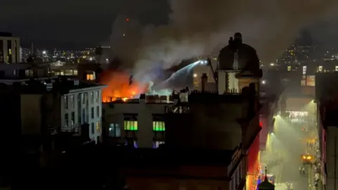 Fire on city centre rooftop with streets below and fire hose trained on the fire from large ladder.