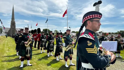 Members of the band who are dressed on green tartan tunics and kilts with white boots and glengarry caps.