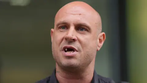 PA Media Jonathan Glover wears a black shirt. He has a shaved head and his mouth is open, revealing that he is missing two teeth on his upper row.