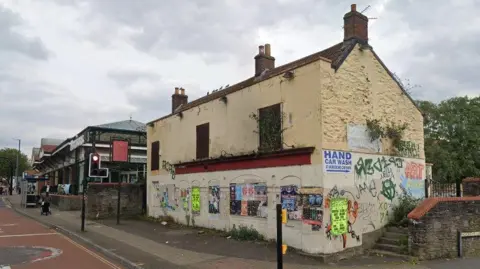 Google An empty, derelict looking building on the corner of a street. It is covered in posters and graffiti.
