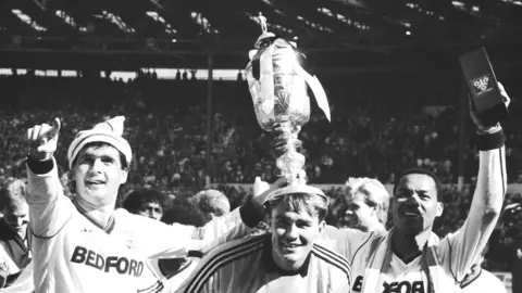 PA Media Luton Town captain Steve Foster (l) and two goal scorer Brian Stein (r) crown their goalkeeper Andy Dibble with the Littlewoods Challenge Cup after beating Arsenal 3-2 in a thrilling final.