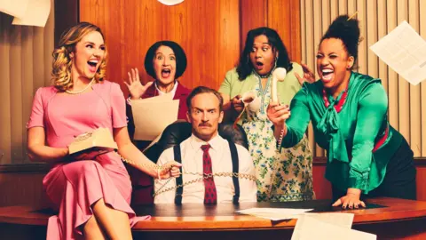 Rebecca Pitt A man wearing a tie and braces sits behind an office desk wrapped in an old-style telephone handset's cable. A woman in a pink dress sits on the desk holding the old rotor-dial phone. Three other women stand behind the desk with one holding the receiver. All are laughing apart from the man, who is frowning.
