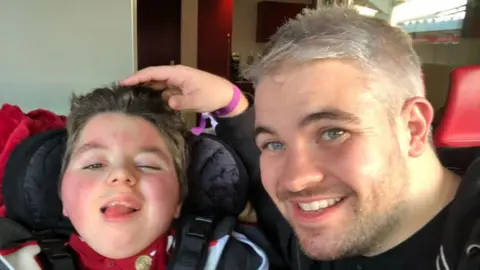 Paul Woodward Paul Woodward is sitting next to his son Kalel with his hand resting on his head. Kalel is sticking his tongue out while Paul is smiling. 