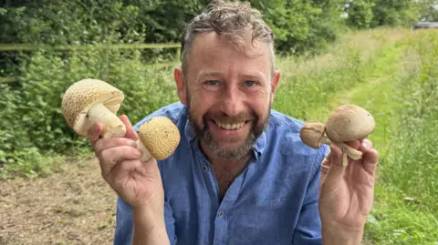 Supplied A man crouched on the ground. He is smiling, wearing a blue shirt, and holding mushrooms in both his hands that he is holding up to each side of his face. There is grass and a hedge behind him.