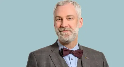 William Alberque Man with gray hair and a gray beard. He is wearing a cheque jacket and bow tie with a blue shirt. He is smiling. Behind him is a blue background.