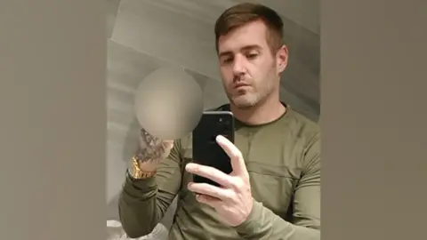 GMP Jamie Rothwell is posing for a selfie in a mirror. He is wearing a khaki top and a gold watch. He is making an offensive gesture with his right hand.